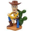 Gartenfigur woody zeigt eine sitzende figur mit braunem cowboyhut, gelb kariertem hemd und blauen hosen auf kaktee