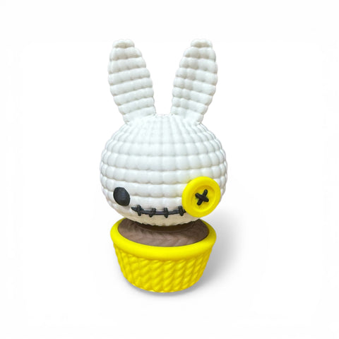 Weißer gehäkelter 3d voodoo-hase mit gelbem knopfauge und gesticktem gesicht in gelbem cupcake-halter sitzt