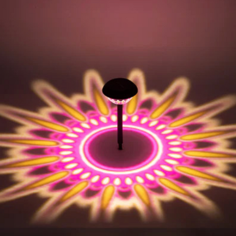 Eine schwarze solar-gartenlampe mit rundem facettiertem kopf wirft ein pink-gelbes 360° muster auf den boden